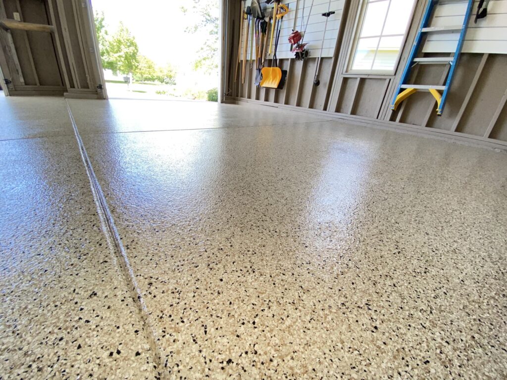 A garage with a newly polished, speckled epoxy floor. Various tools hang on the wall, natural light filtering in from the open door.