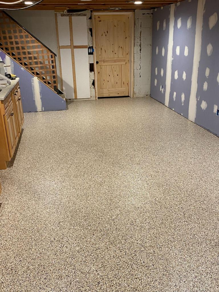 A basement room features a speckled floor, wooden door, partially finished walls, staircase, and kitchen area under exposed ceiling beams.