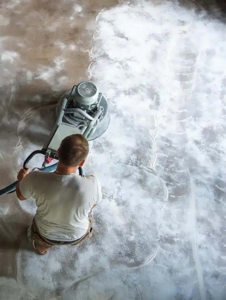 A person operates a floor-cleaning machine, scrubbing a concrete floor in an indoor setting, creating a pattern in the dust and grime.