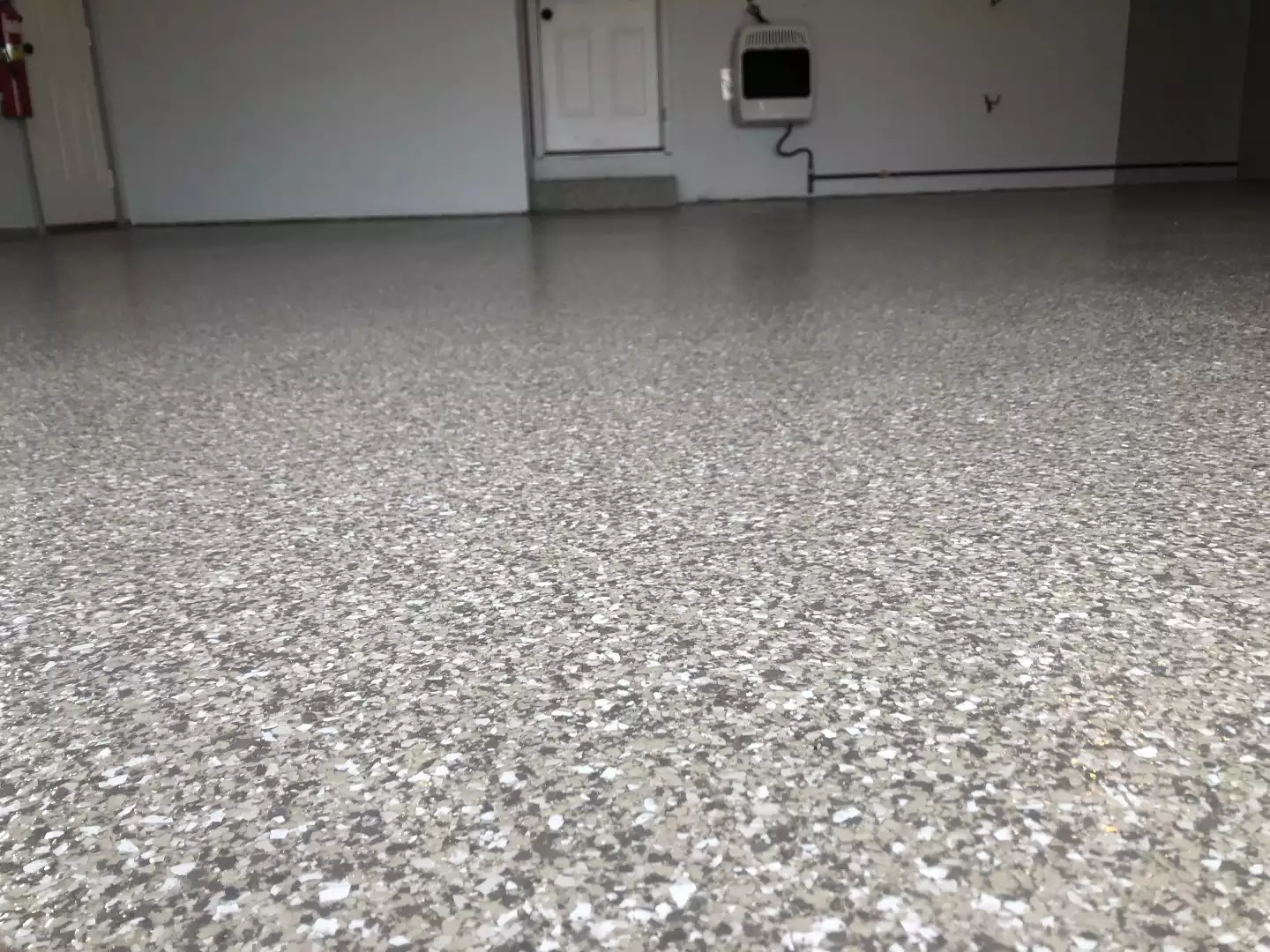 A close-up of a gray speckled floor in a clean, empty room with white walls, a door, and a mounted heater.